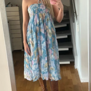 Blommig axelbandslös sommarklänning - Axelbandslös klänning i ljusblått med blommigt mönster i pastellfärger. Smockad över bysten och vidd i kjolen, perfekt för varma dagar. Lätt och luftig känsla, midilängd och utan ärmar.