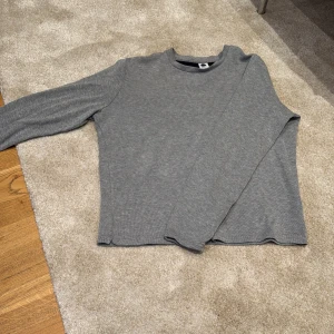 Grå långärmad tröja från NN07 - En stilren grå långärmad tröja från NN07 i storlek L. Tröjan har en rund halsringning och är tillverkad i ett mjukt, ribbat material som ger en clean och avslappnad look. Perfekt för lager-på-lager under höst och vinter.