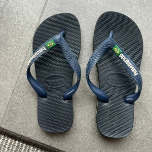 Blå Havaianas flip-flops - Säljer ett par klassiska blå Havaianas flip-flops med brasilianska flaggan på remmen och vit logga. Använda 1-2 gånger Sval och bekväm design med räfflad sula och mjukt syntetmaterial, perfekta för stranden eller sommardagar.