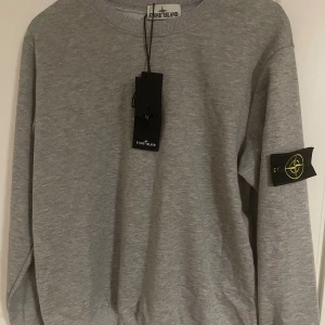 Grå sweatshirt från Stone Island - Snygg grå sweatshirt från Stone Island med klassisk logga på ärmen. Tröjan har rund halsringning, långa ärmar och ribbade muddar. Tillverkad i mjuk bomull för en skön och avslappnad stil. Perfekt för dig som gillar streetwear och stilrena plagg.