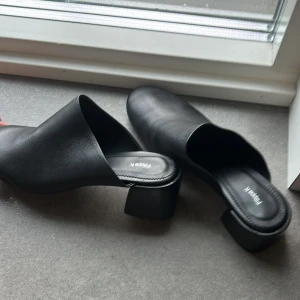 Svarta skinnmules från Filippa K - Stilrena svarta mules från Filippa K i äkta skinn med låg blockklack och rund tå. Skorna har öppen häl och en minimalistisk design som passar till många olika looks. Perfekta för dig som gillar enkel och clean stil.