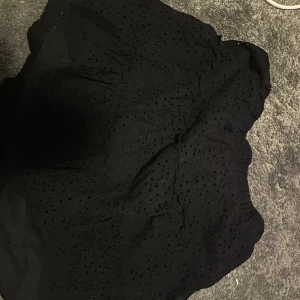 Svart kortkjol med broderade hål - Säljer en svart kortkjol med broderade hål och elastisk midja. Kjolen har en lätt volangig form och är perfekt för dig som gillar en stilren men ändå detaljrik look. 