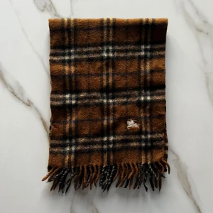 Burberry halsduk - En brun Burberry halsduk med klassiskt rutigt mönster i svart, beige och vitt. Halsduken har fransar i ändarna och är tillverkad i ett mjukt, värmande material som passar perfekt för kyliga dagar. Perfekt accessoar för att lyfta din höst- eller vinterstil. Detta är en mindre modell än de vanliga Burberry halsdukarna!
