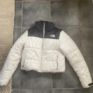 Vit och svart pufferjacka The North Face - Snygg pufferjacka från The North Face i vit och svart med hög krage och dragkedja framtill. Jackan har quiltad design och två fickor med dragkedja. Perfekt för kalla dagar och riktigt trendig look. Använd en vinter så näst intill ny