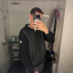 Svart cardigan från Moncler - Svart cardigan från Moncler med quiltad framsida och dragkedja. Cardiganen har två fickor med dragkedja och en klassisk Moncler-logga på ärmen. Materialet är bomull i ärmar och rygg, medan framsidan är dunvadderad för extra värme.