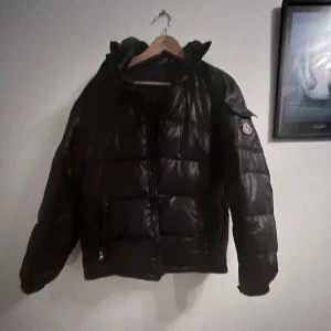 Svart pufferjacka med huva - Säljer en svart pufferjacka. Jackan har huva, dragkedja framtill och två fickor med dragkedja. Den är vadderad och har en glansig finish som ger en riktigt clean look. Perfekt för kalla dagar när du vill hålla stilen. 