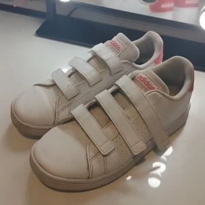 Vita adidas sneakers med kardborre - Säljer ett par vita adidas sneakers i storlek 35 med tre kardborreband och diskreta perforeringar på sidorna. Skorna är väl användna så tänk på att dom är smutsiga men går att ta bort med sko puts🤍