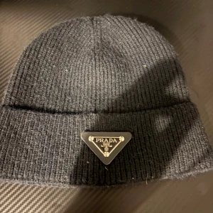 Svart ribbstickad mössa från Prada - Svart ribbstickad mössa från Prada med klassisk uppvikt kant och ikonisk trekantig logotyp i metall framtill. Mössan är tillverkad i mjukt material som håller dig varm och ger en clean och lyxig look. Perfekt accessoar för att lyfta din outfit.