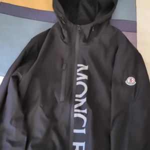 Svart vindjacka från Moncler med huva - Säljer en svart vindjacka från Moncler med stor logga framtill och klassisk patch på ärmen. Jackan har huva, dragkedja och en bröstficka med zip. Perfekt för dig som gillar stilrena och sportiga plagg. Den är vattentät och har jätte bra kvalité. Kan fraktas eller mötas upp I Stockholm 