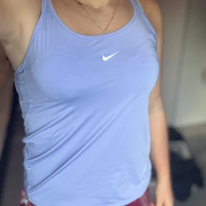 Ljuslila linnetopp från Nike - Säljer en ljuslila linnetopp från Nike med smala axelband och klassisk vit swoosh-logga framtill. Toppen är i ett mjukt och stretchigt syntetmaterial, perfekt för träning eller chill. Snygg och enkel design som är lätt att matcha.