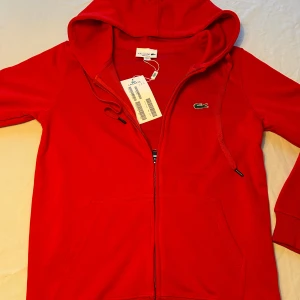 Röd  Lacoste hoodie - Riktigt snygg röd hoodie  Lacoste med klassisk krokodillogga på bröstet. Tröjan har huva med snörning, dragkedja framtill och kängurufickor. Tillverkad i mjuk bomullsmix som känns skön mot huden. Perfekt för en chill och sportig stil.