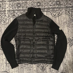 Svart Moncler Cardigan  - Svart Moncler cardigan med stickade ärmar och quiltad front. Dragkedja framtill och två dragkedjeförsedda fickor. Klassisk Moncler-logga på ena ärmen. Perfekt för höst och vinter när du vill hålla stilen. Och man kan även skanna loggan på sidan. Priset är ej hugget i sten så var inte rädda att ställa frågor 😁
