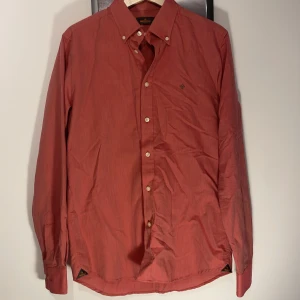 Röd skjorta från Morris  - Snygg röd skjorta från Morris i storlek M, med klassisk button down-krage och broderad logga på bröstet. Skjortan har vita knappar och är tillverkad i ett mjukt bomullstyg. Perfekt för dig som gillar stilrena detaljer.
