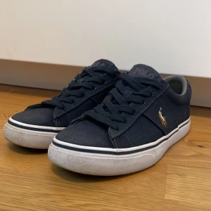Mörkblå sneakers från Polo Ralph Lauren - Snygga mörkblå sneakers från Polo Ralph Lauren med klassisk låg profil och vit platt sula. Skorna har snörning och är tillverkade i slitstark bomullstextil. På sidan finns Polo-loggan broderad i brunt och vitt. Perfekta för en chill och stilren look.
