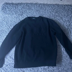 Svart sweatshirt från Hugo Boss  - Svart sweatshirt från Hugo Boss i storlek XL med stort broderat BOSS-logo framtill. Tröjan har rund halsringning och långa ärmar, tillverkad i mjuk bomull för en clean och stilren look.