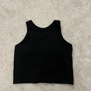 Svart ribbad linnetopp från H&M Sport - Svart ribbad linnetopp från H&M Sport i storlek M. Toppen har en klassisk rund halsringning och är ärmlös med breda axelband. Perfekt för träning eller chill dagar. Materialet är mjukt och stretchigt för extra komfort.