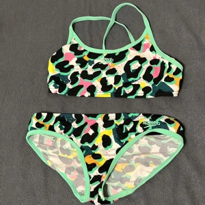 Leopardmönstrad bikini från Speedo - Cool bikini från Speedo med leopardmönster i svart, grön, gul, rosa och vit. Toppen har smala korsade axelband och gröna kanter, medan trosan har samma fräscha färgdetaljer och gröna kant. Perfekt för dig som vill sticka ut på stranden eller vid poolen.