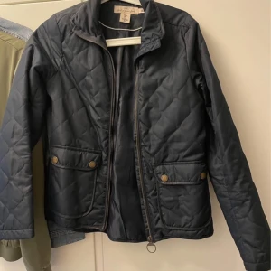 Svart quiltad jacka från Lexington - Quiltad svart jacka från Lexington med dragkedja och två stora fickor framtill med tryckknappar. Jackan har ståkrage och klassisk passform, perfekt för kyliga dagar. Snygg och stilren design som funkar till det mesta.