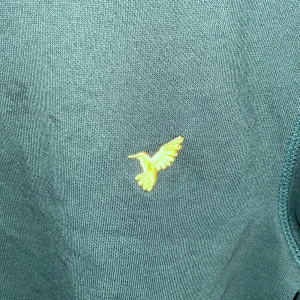 Mörkgrön sweatshirt från Pier One - Mörkgrön sweatshirt från Pier One med rund hals och lång ärm. På bröstet finns en gul broderad fågel som detalj. Tröjan är tillverkad i mjuk bomull och har ribbade muddar vid ärmslut och nederkant. Perfekt för chill dagar.