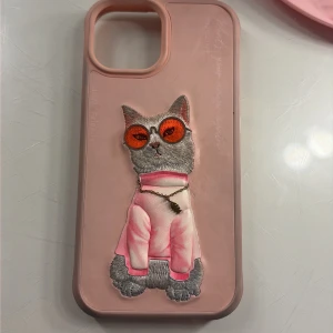 Nimmy x NIMMY Friends Cat iPhone Case. iPhone 15 - Snygg rosa iPhone-skal från Nimmy x NIMMY Friends med broderad katt i solglasögon och rosa tröja. Skalet är i fint skick utan synligt slitage och ger bra skydd till din mobil. Perfekt för dig som vill ha ett unikt och trendigt mobilskal.