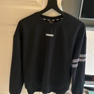 Svart sweatshirt från Stronger XS - Svart nästan oanvänd sweatshirt från Stronger med vit logga på bröstet och två vita randiga detaljer på ena ärmen. Klassisk rund halsringning och lång ärm, perfekt för en sportig och avslappnad stil.