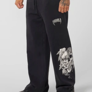 Svarta mjukisbyxor med dödskalletryck - Svarta mjukisbyxor från YoungLA med bred passform och coolt vitt tryck av dödskalle, rosor och orm på benen. Byxorna har snörning i midjan och en bakficka. Aldrig använda och prislapp kvar 