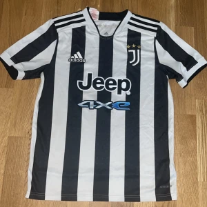 Juventus randig matchtröja Adidas - Juventus officiella fotbollströja från Adidas med klassiska svarta och vita ränder, klubbmärke med tre stjärnor och Jeep-sponsring på bröstet. Tröjan har korta ärmar, rund hals och är tillverkad i lätt funktionsmaterial.