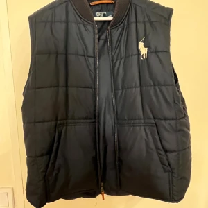 Mörkblå dunväst från Polo Ralph Lauren XL - Svart quiltad dunväst från Polo Ralph Lauren i storlek XL. Västen har dragkedja framtill, ribbad krage och klassisk Polo-logga broderad på bröstet. Tillverkad i 100% polyester och har ett stilrent rutmönster över hela västen. Passar bra nu hösten och en bra fars dag present (9 nov) eller till dig själv.