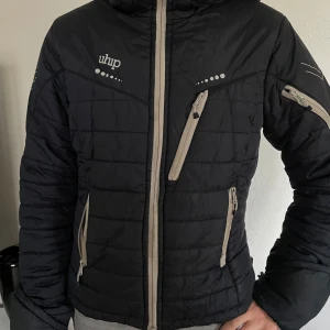 Svart quiltad pufferjacka från Uhip - Säljer en svart pufferjacka från Uhip med grå dragkedjor och avtagbar huva. Jackan har flera praktiska fickor med dragkedja och ett quiltat mönster. Perfekt för kyliga dagar och enkel att matcha med olika outfits. Lite sliten längst ner i ärmarna och en lagning i vänster ficka 