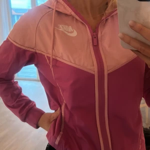 Rosa Nike vindjacka med huva - Nike vindjacka i två nyanser av rosa med vit logga på bröstet. Jackan har huva med snörning, hel dragkedja framtill och elastiska muddar. Lätt och sportig design, perfekt för utomhusaktiviteter. Snygga detaljer och skön passform.