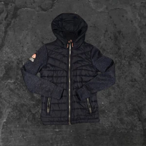 Superdry cardigan  - Säljer en svart hybridjacka från Superdry Mountain med quiltad front och huva. Jackan har dragkedja, två fickor med dragkedja och broderad logga på ärmen. Ärmar och rygg är i mjukare material för extra komfort. Perfekt för kyliga dagar.