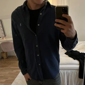 Blå skjorta från Ralph Lauren - Säljer en klassisk blå skjorta från Ralph Lauren i modellen Featherweight Mesh. Skjortan har lång ärm, knappar framtill och ikoniska broderade loggan på bröstet. Tillverkad i ett lätt och luftigt bomullsmaterial som är perfekt för en stilren look. Tveka inte att fråga vid minsta fundering!