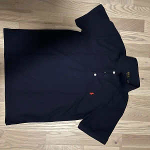 Mörkblå pikétröja från Ralph Lauren - Klassisk mörkblå pikétröja från Ralph Lauren med röd broderad logga på bröstet. Tröjan har korta ärmar, krage och knappar framtill. Perfekt för dig som gillar stilrena och tidlösa plagg med en sportig touch.