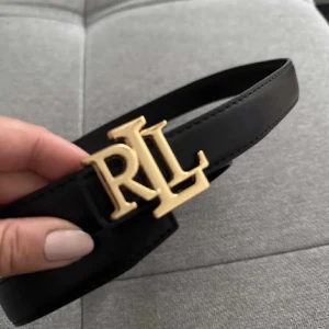 Svart skärp från Ralph Lauren - Snyggt svart bälte från Ralph Lauren med en stor RL-logga i guld som spänne. Bältet är gjort i slätt skinn och har en klassisk, stilren design som passar till jeans eller kostymbyxor. Perfekt accessoar för att lyfta din outfit.