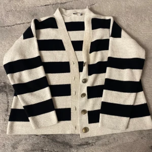 Randig kofta med knappar i svart och vitt - Snygg kofta med breda svarta och vita ränder och fem metallknappar framtill. Tröjan har lång ärm och är stickad i ett mjukt material.  Kostade: 200