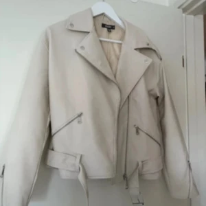 Beige skinnjacka  - Snygg beige skinnjacka från Missguided med klassisk bikerstil. Jackan har bred krage, silverfärgade dragkedjor, bälte i midjan och långa ärmar med zip-detaljer. Perfekt för dig som gillar en clean och cool look.