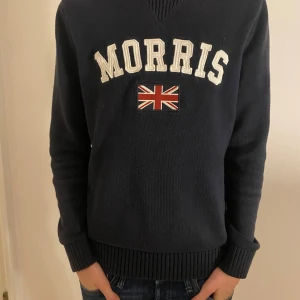 Mörkblå stickad tröja Morris - Snygg mörkblå stickad tröja från Morris med vit broderad logga och en Union Jack-flagga på bröstet. Tröjan har ribbade muddar vid ärmslut och nederkant samt rundad hals med diskret v-detalj. Perfekt för dig som gillar klassisk och sportig stil. Modellen på bilden är 176cm lång