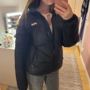 Svart pufferjacka från Fila - Svart pufferjacka från Fila med broderad logga på bröstet. Jackan har hög krage, dragkedja framtill och två sidofickor med dragkedja. Perfekt för kalla dagar och har en snygg, enkel design som funkar till jeans eller hoodies. Använd få gånger