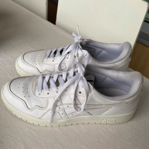 Vita Asics sneakers i storlek 38 - Fräscha vita Asics sneakers med klassisk låg profil och rund tå. Ovandel med perforerade detaljer och  Asics-loggor på hälen och innersulan. Skorna har snörning och en platt gummisula.