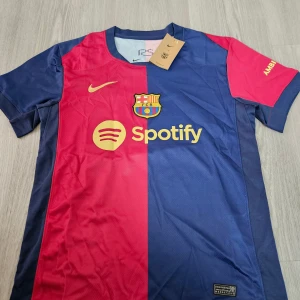 Barça Messi 10 Nike fotbollströja - FC Barcelona fotbollströja med Messi 10-tryck, officiell Nike-modell. Tröjan är blå och röd med gula detaljer, klubbmärke och sponsorer som Spotify och UNHCR. Tillverkad i lätt och ventilerande material, perfekt för match eller träning.