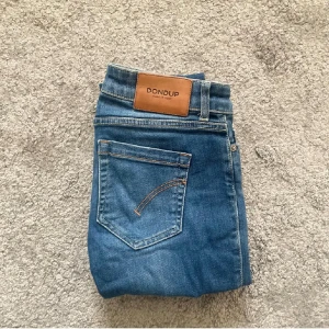 Blå jeans från Dondup - Snygga blå jeans från Dondup med klassisk femficksdesign och bruna sömmar. Modellen har rak passform och normal midja, med en stilren läderpatch bak i midjan. Tillverkade i slitstarkt denim-material. Perfekta för dig som gillar italiensk design.När jag köpte de så köpte jag de som storlek S. Mått 90 cm i längd och 35 cm Waist