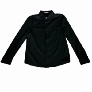Svart overshirt från Zara - Stilren svart overshirt från Zara med klassisk krage, två bröstfickor och knäppning framtill. Skjortan har lång ärm och en rak passform, perfekt att bära som lager över t-shirt. Materialet känns mjukt och följsamt.