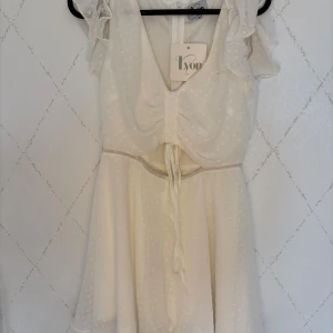 Vit prickig kortklänning från Lyon - Supersöt vit kortklänning från Lyon med diskreta prickar, volangärmar och snörning framtill. Klänningen har v-ringning, markerad midja och är tillverkad i ett lätt, luftigt material som känns perfekt för varma dagar.