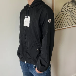 Svart vindjacka från Moncler med huva - Svart vindjacka från Moncler med huva och dragkedja framtill. Jackan har två snedställda fickor med tryckknappar och en diskret Moncler-logga på ärmen. Lätt material i polyester och snygga detaljer med röd och vit rand på huvan.