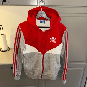 Adidas röd och grå hoodie med dragkedja - Snygg hoodie från Adidas i rött, grått och vitt med klassiska ränder på ärmarna. Tröjan har huva med snörning, dragkedja framtill och Adidas-logga broderad på bröstet. Tillverkad i mjuk bomull och polyesterblandning, perfekt för chill eller träning.