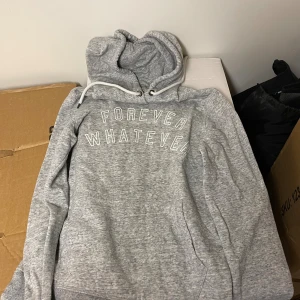 Grå hoodie med tryck från L.O.G.G. - Gråmelerad hoodie från L.O.G.G. med vit text 'FOREVER WHATEVER' på bröstet. Tröjan har huva med snörning och en klassisk känguruficka framtill. Mjuk och skön bomullsmix, perfekt för chill dagar. Passar dig som gillar avslappnad stil.