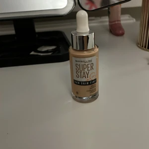 Maybelline Super Stay 24H Skin Tint 10 - Maybelline Super Stay 24H Skin Tint i nyansen 10. Flytande foundation med pipettapplikator, ger en naturlig finish och lång hållbarhet. Kommer i en glasflaska med silverfärgat lock. Perfekt för dig som vill ha en lätt och jämn bas. Köpt för 120 och aldrig använd förutom att testa. Säljer då den är i fel färg för mig