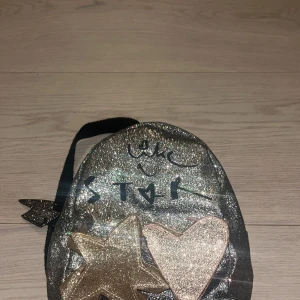 Glittrig silver ryggsäck med stjärna och hjärta - Snygg liten ryggsäck i glittrigt silver med svarta axelband. Framtill finns en guldig stjärna och ett hjärta i glitter, samt texten 'like a star'. Väskan har dragkedja och är perfekt för barn som gillar att sticka ut med coola detaljer.