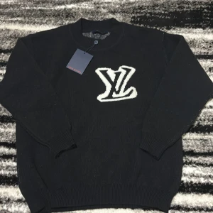 Svart stickad tröja Louis Vuitton - Svart stickad tröja från Louis Vuitton med stort vitt LV-logo på bröstet och ryggen. Klassisk rund halsringning och ribbade muddar vid ärmslut och nederkant. Perfekt för dig som vill ha en lyxig och stilren look.