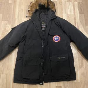 Svart Canada Goose Expedition Parka - Canada Goose Expedition Parka i svart med ikonisk röd och blå logga på bröstet. Jackan har stor huva med pälsimitation, flera stora fickor och tydlig EXPEDITION-detalj på ryggen. Perfekt för kalla vinterdagar och riktigt snygg streetstil. Dragkedjan funkar inte 